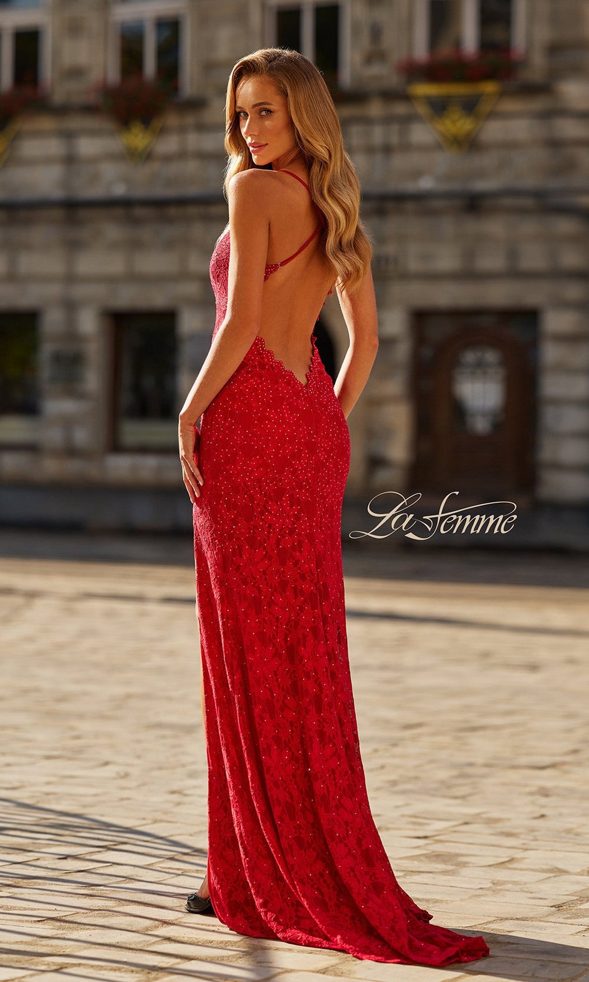 A woman poses outdoors in the Long Prom Dress: La Femme 33647, a floor-length red gown with lace details, thin straps, and an open back. The La Femme logo appears at the bottom of the image.